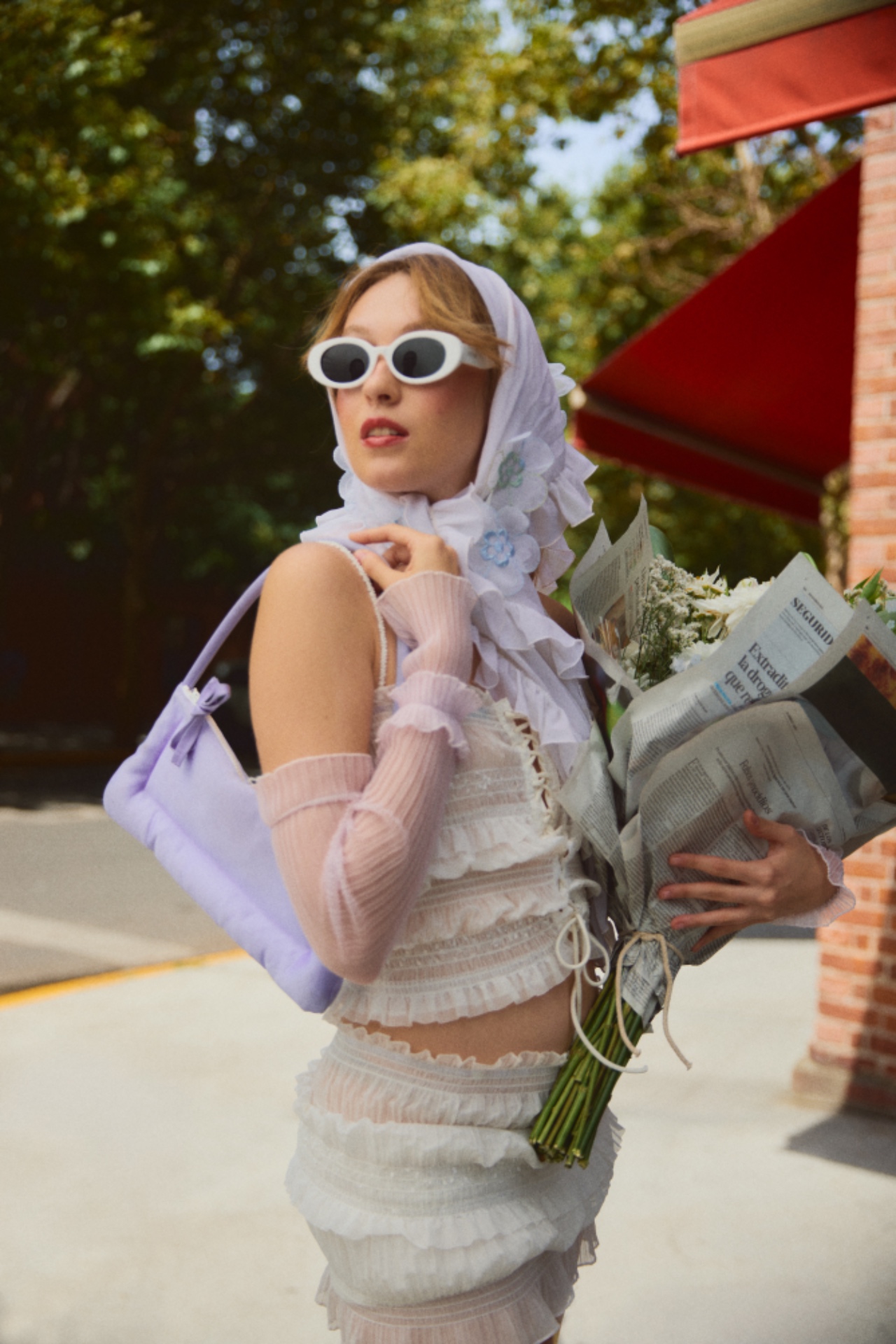 Woman in white sunglasses and a white headscarf carrying flowers and a newspaper with a pale lavender bag