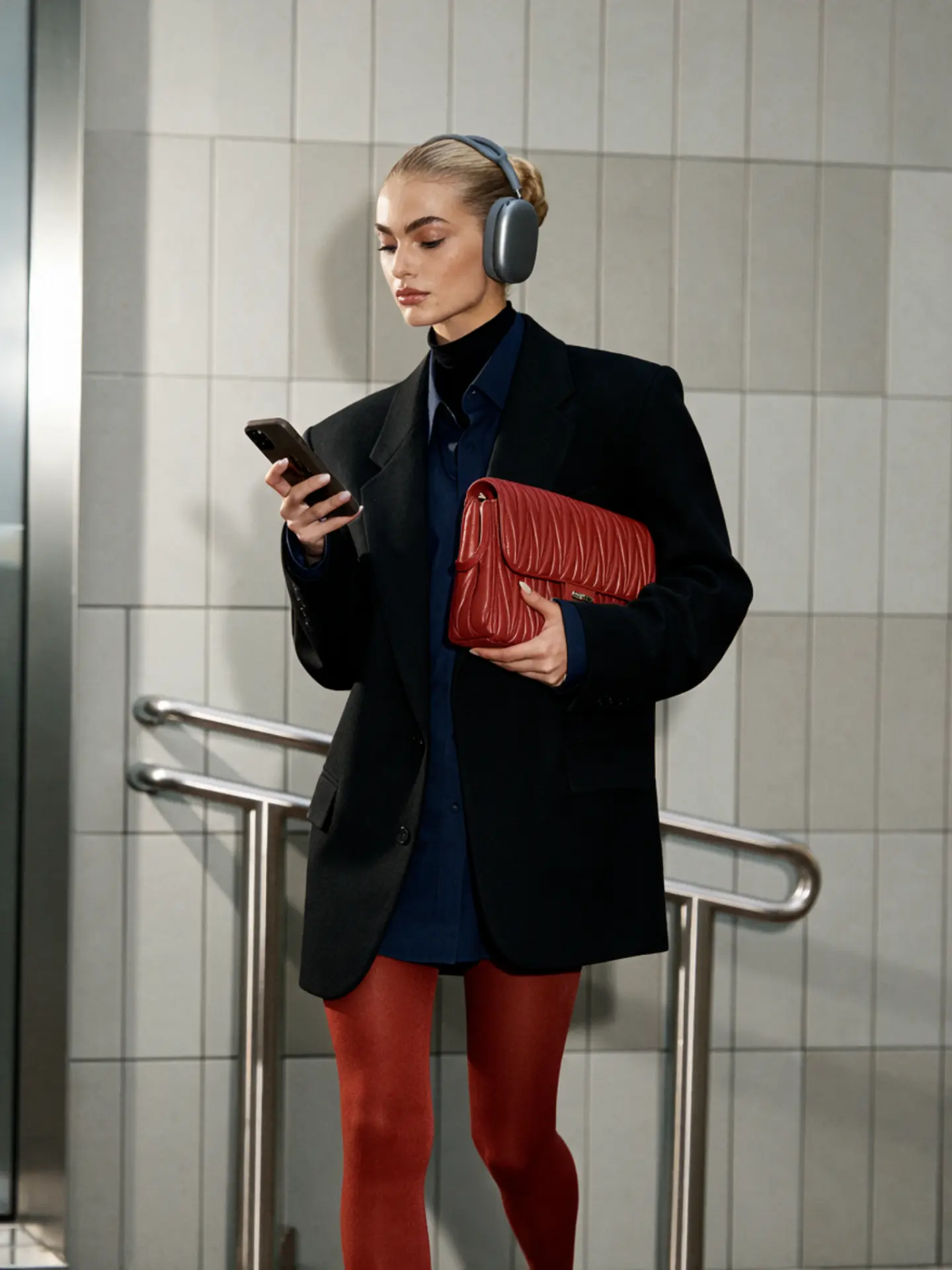 Woman wearing headphones and a dark blazer, holding a red bag and looking at her phone beside a neutral tiled wall
