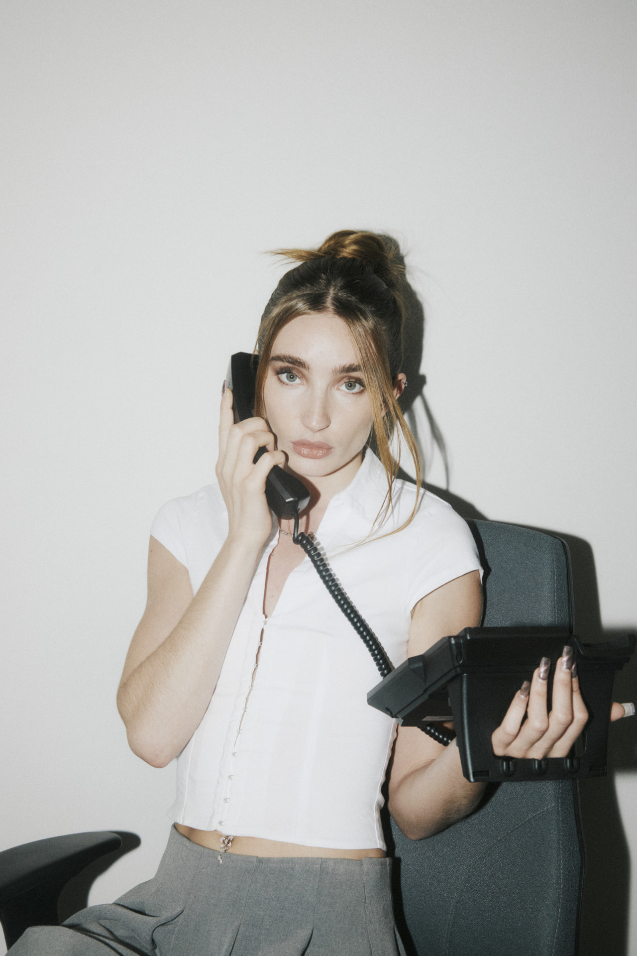 Woman in a white top and grey trousers seated in an office chair holding a telephone receiver and black device